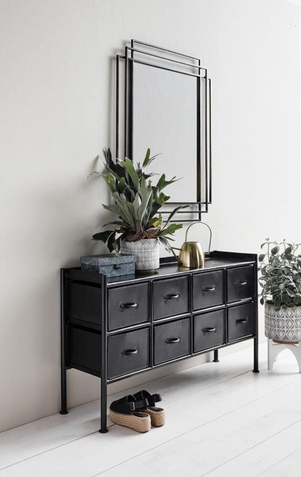 Et elegant sort konsolbord med flere skuffer står op ad en hvid væg og viser TomorrowsDesign, toppet med et spejl, en plante i en keramisk potte og dekorative genstande. Et par hjemmesko sidder på gulvet ved siden af.