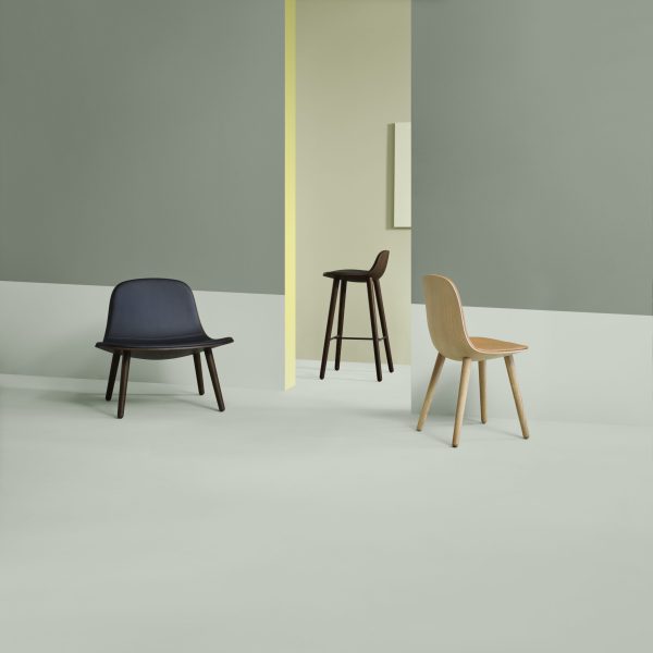Tre moderne stole i et rum med grå og gule vægge har en sort loungestol, en træskammel og en lys træstol fra TomorrowsDesign, med afstand fra hinanden på et lysegråt gulv.