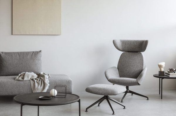 En minimalistisk stue med en grå sofa med et tæppe og pude, en moderne stol med en matchende skammel og et rundt sort sofabord med bøger og pynteting. Et almindeligt lærred hænger på væggen, der fremhæver rummets design.