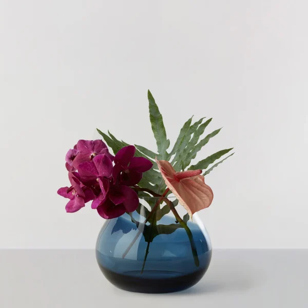En rund, blå RO KOLLEKTION - Blomstervase nr. 23 på en almindelig overflade, indeholdende et arrangement af lyse lyserøde og laksefarvede blomster med grønne, strukturerede blade, mod en lysegrå baggrund.