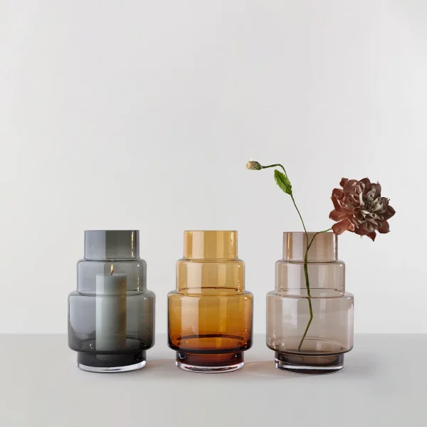 Et arrangement af fem RO COLLECTION - Hurricane No. 69 glasvaser i forskellige nuancer og størrelser, fra transparent til rav til grå, med to med enkelte blomster og en med et tændt stearinlys, vist på en hvid baggrund.