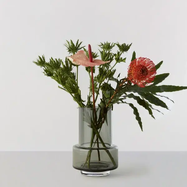 Et simpelt arrangement af eksotiske blomster inklusive en pink anthurium og en rød protea, sat i en RO COLLECTION - Hurricane No. 26 vase mod en almindelig, lysegrå baggrund.