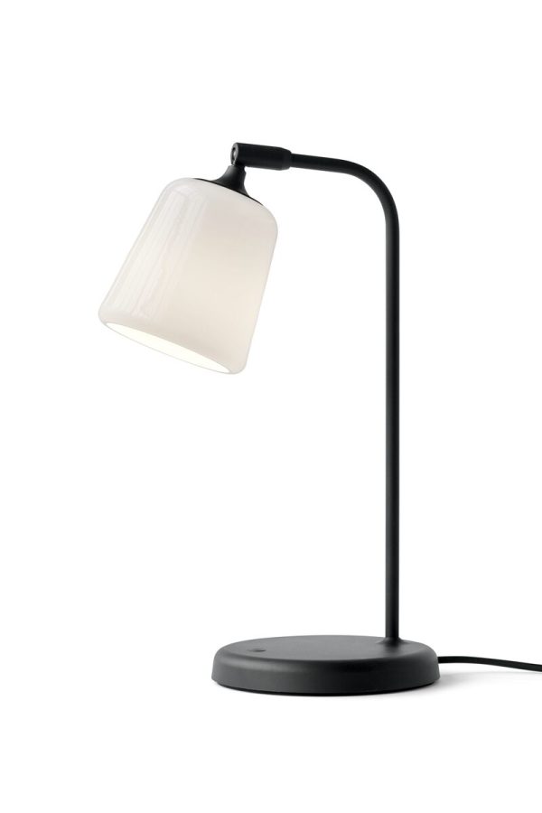 Et moderne NYT VÆRK - Materiale Bordlampe med sort stativ og en gennemskinnelig hvid lampeskærm, oplyst og sat mod en almindelig hvid baggrund.