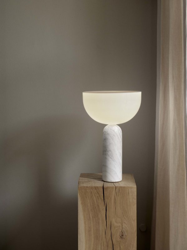 En moderne lampe med en cirkulær, uigennemsigtig skærm sidder oven på en hvid marmorbase, som er placeret på en træsokkel i et rum med bløde, neutrale vægge. Lyset kaster en blid skygge på baggrunden, der perfekt komplementerer interiørtemaet i TomorrowsDesign.