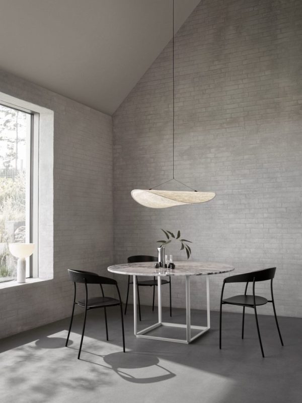 En minimalistisk spiseplads med et rundt marmorbord og to sorte stole under en unik oval pendel. Værelset har designmøbler grå murstensvægge og et stort vindue med udsigt til grønt udenfor.