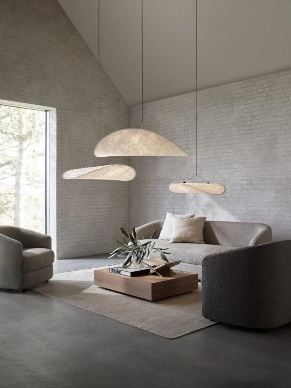 En minimalistisk stue med neutrale toner med to overdimensionerede pendellamper over et NYT VÆRK - Massivt sofabord, omgivet af bløde, afrundede lænestole og store vinduer, der slipper naturligt lys ind.