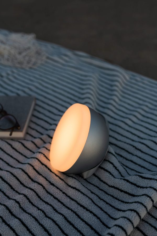 ET NYT VÆRKER - Sphere Adventure Lampe lyser orange på et stribet tæppe ved siden af en bog og briller i tusmørket, hvilket skaber en hyggelig atmosfære.