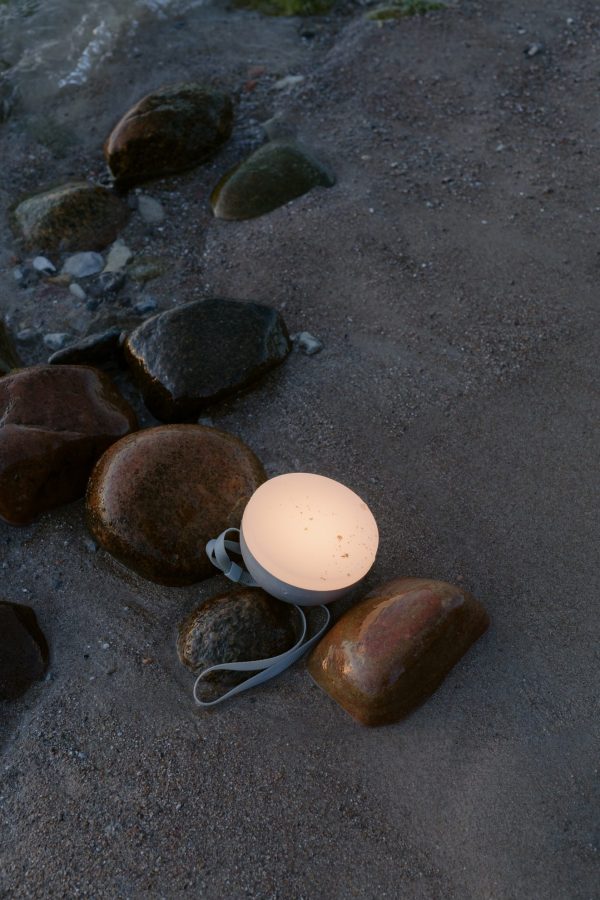 Et hvidt NYT VÆRK - Sphere Adventure Lampe, der udsender et blødt skær, sidder på en sandstrand omgivet af store, våde sten i skumringen. Lampen er sat i stikkontakten, med ledningen trukket ud af rammen.