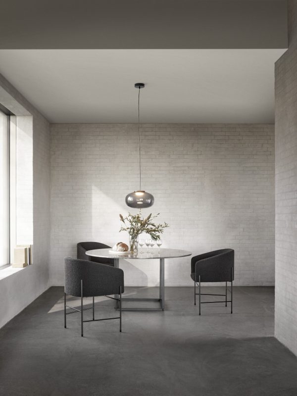 En minimalistisk spiseplads med et rundt bord med to mørke polstrede stole, under et NYT VÆRK - Karl-Johan Pendel Ø40. Rummet har grålige murstensvægge og betongulv, med naturligt lysindfald fra et vindue.