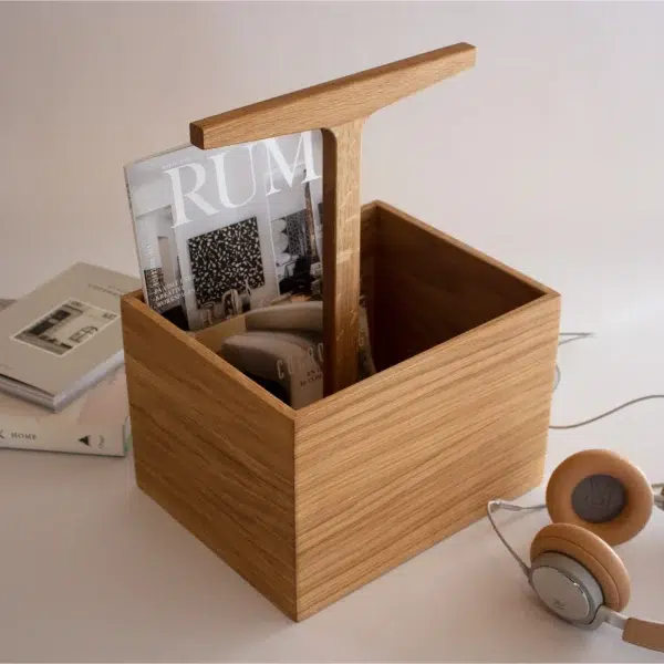 En magasinholder i træ med en lys egetræsfinish, med et minimalistisk design, formet som en kasse med en forlænget arm, der rummer flere magasiner. Ved siden af, på en hvid overflade, står et par høretelefoner og en stak bøger.