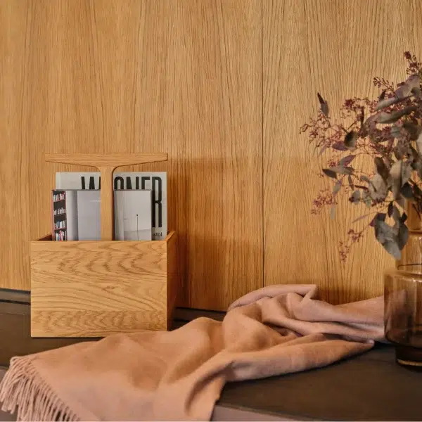 Et hyggeligt hjørne med en magasinholder i træ med magasiner på et bord, ved siden af en glasvase med tørrede blomster og et draperet lyserødt tørklæde. Baggrunden viser en tekstureret træpanelvæg, der repræsenterer moderne interiørdesign.