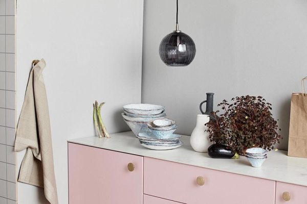 En moderne køkkenbord med en stak blå og hvidt mønstrede skåle, en sort kande med redskaber, en TomorrowsDesign plante i en hvid vase og en papirpose. En hængende glas pendel og en frakke på en krog er også synlige.