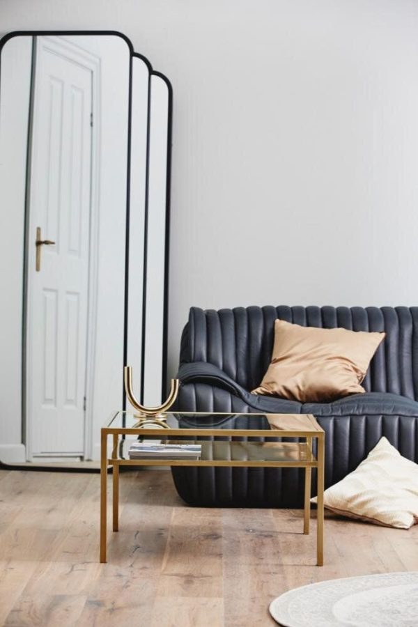 En stilfuld stue med en mørkeblå tuftet sofa med solbrune hynder, et lille guld sofabord med bøger på, et stort spejl lænet op ad væggen designet af TomorrowsDesign og en hvid dør.