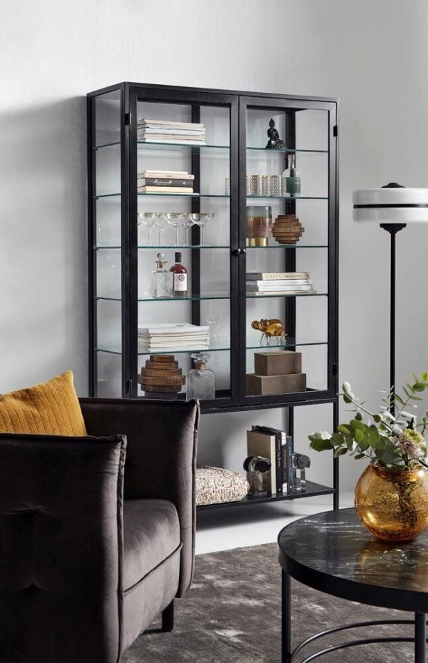 Et moderne vitrineskab i glas med en sort metalramme, med forskellige dekorative genstande og bøger. Den står op ad en lysegrå væg nær en mørk fløjlsstol og rundt, sort sofabord fra TomorrowsDesign.