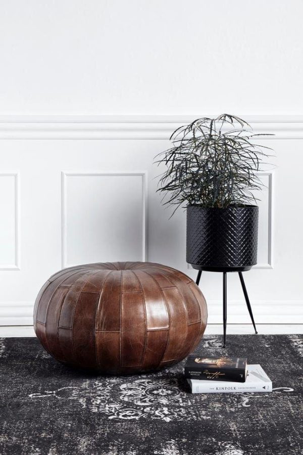 Et stilfuldt indvendigt setup med en læderpuf og en høj, sort metalplantekasse, der holder en frodig grøn plante ved siden af en stak bøger på et mønstret tæppe, mod en hvid panelvæg. Det sofistikerede interiør omfatter moderne møbler, der komplementerer den overordnede æstetik.