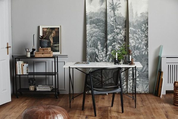 Et pænt indrettet hjemmekontor med et sort metalbord med en stol fra eksklusive designmøbler, sat op ad en væg prydet med et frodigt, skov-tema gobelin. Rummet suppleres af en grå hylde, forskellige planter og kunstnerisk indretning.