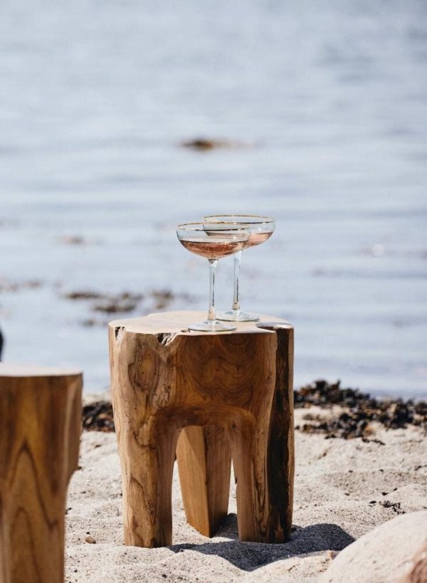 Et glas rosévin placeret på en rustik trætaburet fra TomorrowsDesign ved kanten af en sandstrand med stille blåt havvand og tang i baggrunden.