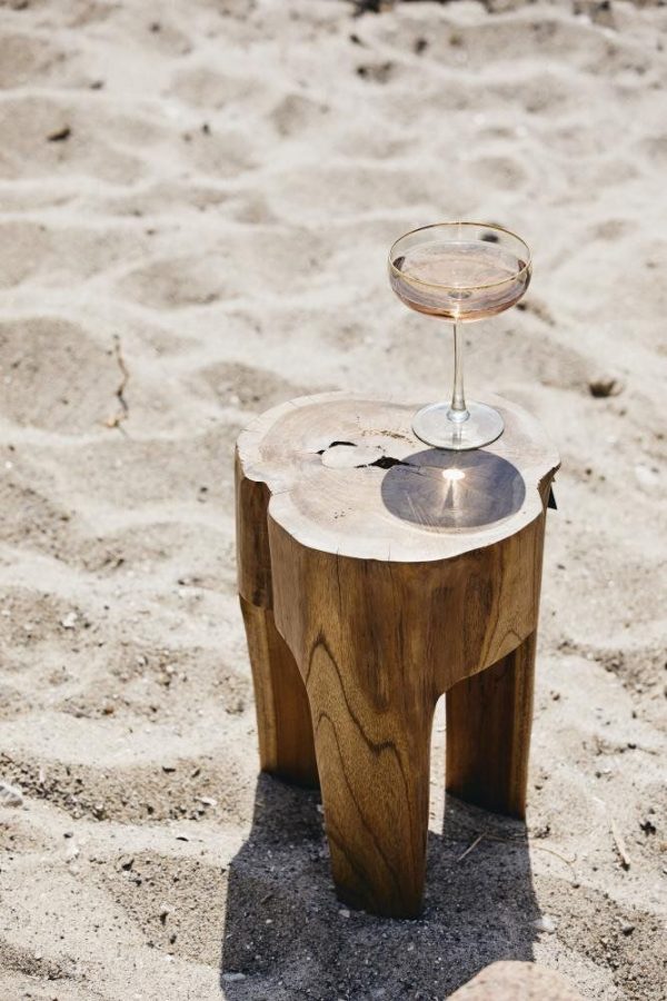 Et glas hvidvin sidder på toppen af en rustik træstub på en sandstrand under stærkt sollys. Stubben, der repræsenterer unikt interiørdesign, bruges som et interimistisk bord.