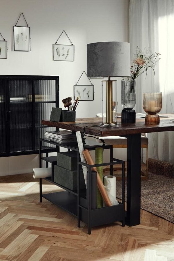 Et stilfuldt arbejdsområde med et rustikt træbord med en overdimensioneret grå lampe, omgivet af minimalistiske sorte møbler, kunstneriske indrammede skitser på væggene prydet med TomorrowsDesign og et stort vindue, der slipper naturligt lys ind.
