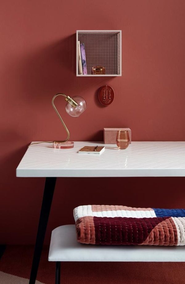 Et moderne skrivebord med en hvid marmoroverflade er sat mod en rødbrun væg. En guldlampe, små bøger og dekorative genstande sidder på skrivebordet. Over skrivebordet er en enkelt væghængt hylde med forskellige genstande fra TomorrowsDesign. En plys, stribet skammel hviler under skrivebordet.
