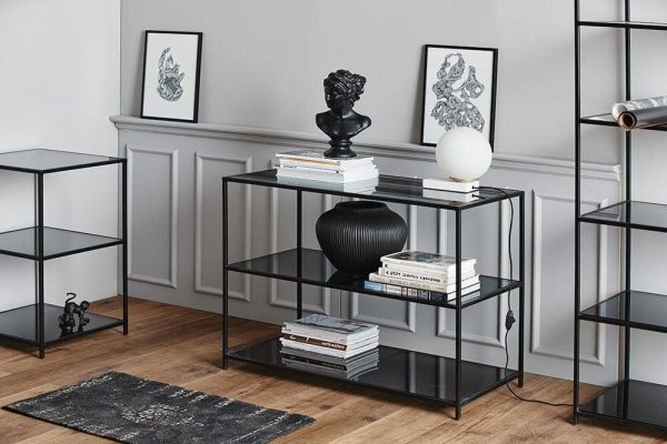 Et pænt organiseret stuehjørne med et moderne sofabord i sort metal og glas prydet med bøger, en sort vase og en hvid kugleformet genstand. Designmøbler og ekstra reoler fremhæver den stilrene, minimalistiske indretning.