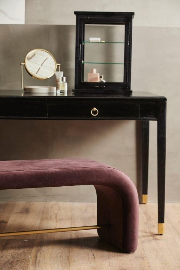 Et elegant sort konsolbord med en lille, åben makeup-boks og et rundt, guldtrimmet ur. En bordeaux fløjlsbænk med guldfødder fra TomorrowsDesign er placeret foran bordet mod en beige væg.