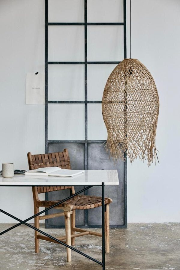 Et minimalistisk arbejdsområde med et elegant hvidt skrivebord med åbne bøger, en vævet rattanstol og en kunstnerisk vævet pendel, der hænger ved siden af et højt vindue med sorte rammer. Interiøret indeholder designmøbler, der komplementerer den overordnede æstetik.