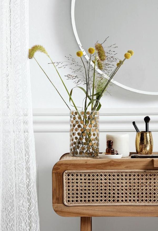 Et moderne boliginteriør med et konsolbord af træ med designmøbler rattandetaljer. På toppen rummer en dekorativ vase vilde blomster ved siden af et hvidt stearinlys og gyldne redskaber i en holder nær et rent hvidt gardin.