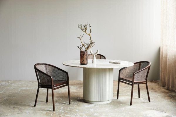 En minimalistisk spisestue med et hvidt rundt bord med en lodret riflet base og tre rattanstole i et rum med nøgne betongulve og gennemsigtige gardiner. En vase med grene og en bog pryder bordet og indkapsler TomorrowsDesign-æstetik.