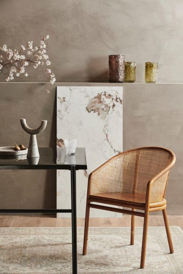 Et moderne, minimalistisk interiørhjørne med en rattanstol ved siden af et sort bord med dekorative genstande, mod en tekstureret beige væg. En marmorplade og glasvaser er også synlige.