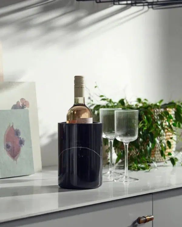 En NORDAL - Marbi Vinkøler af hvidvin ved siden af to klare vinglas på en køkkenbord, med sollys, der kaster skygger og planter i baggrunden.