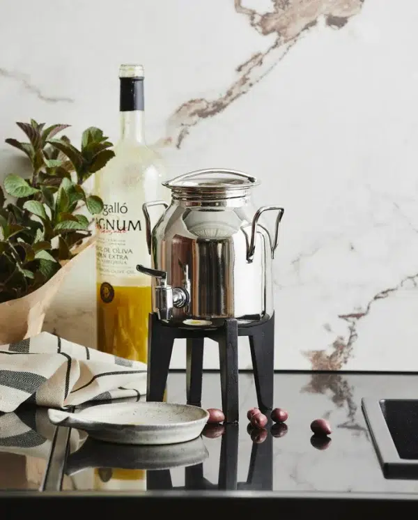 En kaffekande i rustfrit stål står på en lille sort skammel på en marmorbordplade, omgivet af en flaske olivenolie, urter, forskellige redskaber fra TomorrowsDesign og nogle spredte kaffebønner.