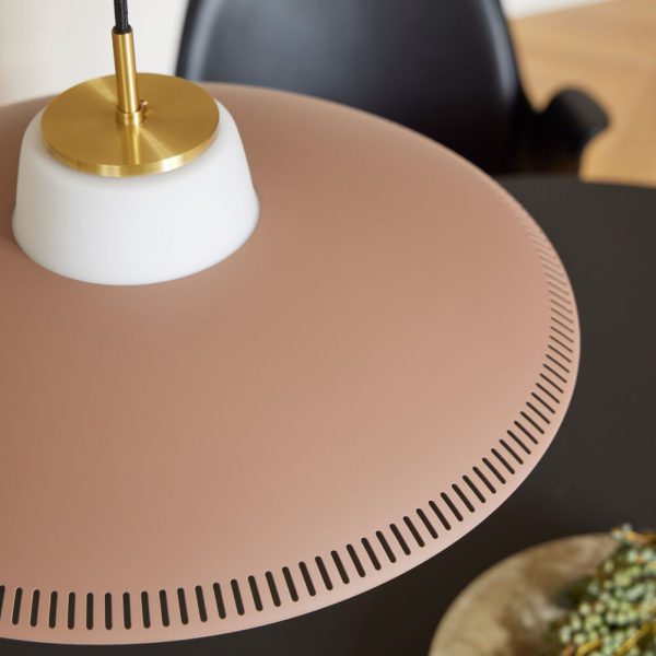 Nærbillede af en moderne pink pendel med guldaccenter, hængende over et sort bord i et subtilt designet rum. Lampen har små ventilationsåbninger rundt om dens nederste kant.