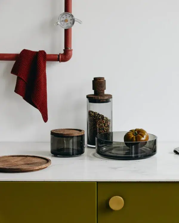 En pænt organiseret køkkenbordplade med NORDAL - Basil Opbevaringsglas, en messingvægt og et grønt skab med en gul knop, fremhævet af et hængende rødt håndklæde.