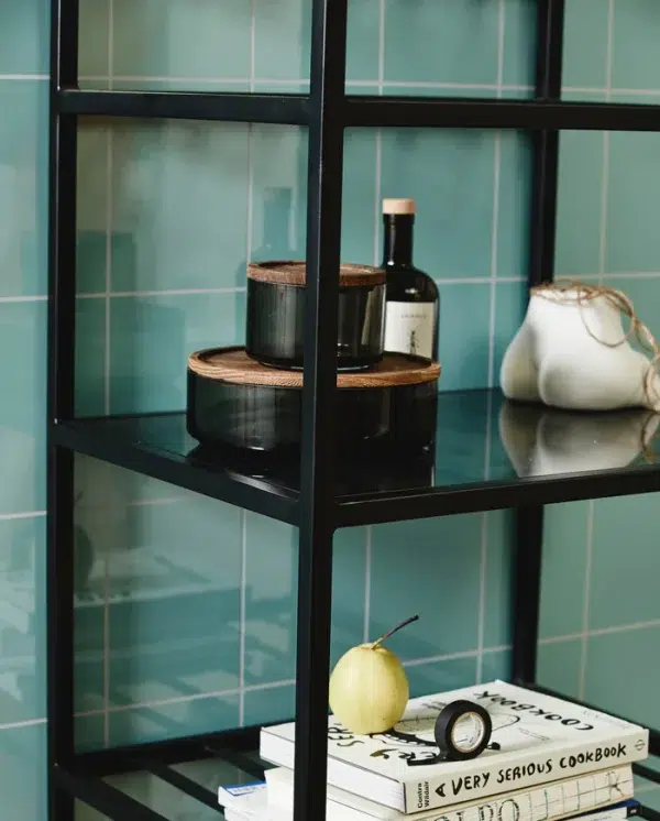 En stilfuld køkkenhylde mod en turkis flisebelagt væg med en stak kogebøger, en flaske vin, to brune beholdere på en træbakke og et NORDAL - Basil Opbevaringsglas med sejlgarn.