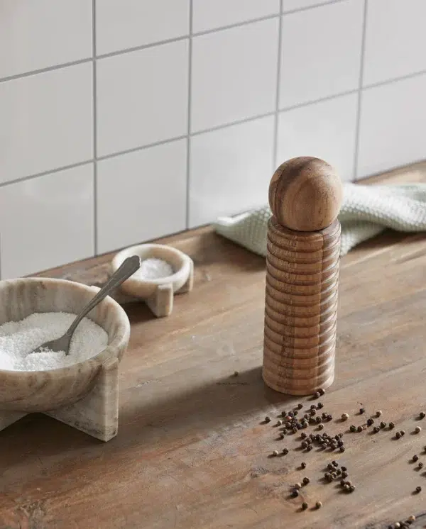 En peberkværn i træ står ved siden af en marmorskål med salt med en ske, på en køkkenbord af træ spredt med peberkorn. En bagplade med hvide fliser og en grøn klud er også synlige, hvilket forbedrer det overordnede design.