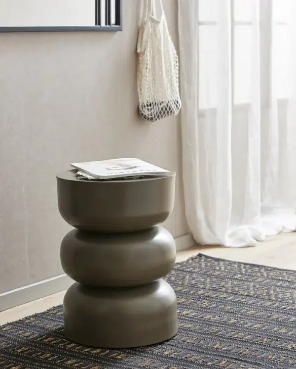 Et moderne, stablet, tre-etageret, mørkegrå sidebord står op ad en beige væg og holder en bog og et par briller. Et NORDAL - Farah Gulvtæppe 250x100 hænger i nærheden, og derunder ligger et mønstret mørkt tæppe.