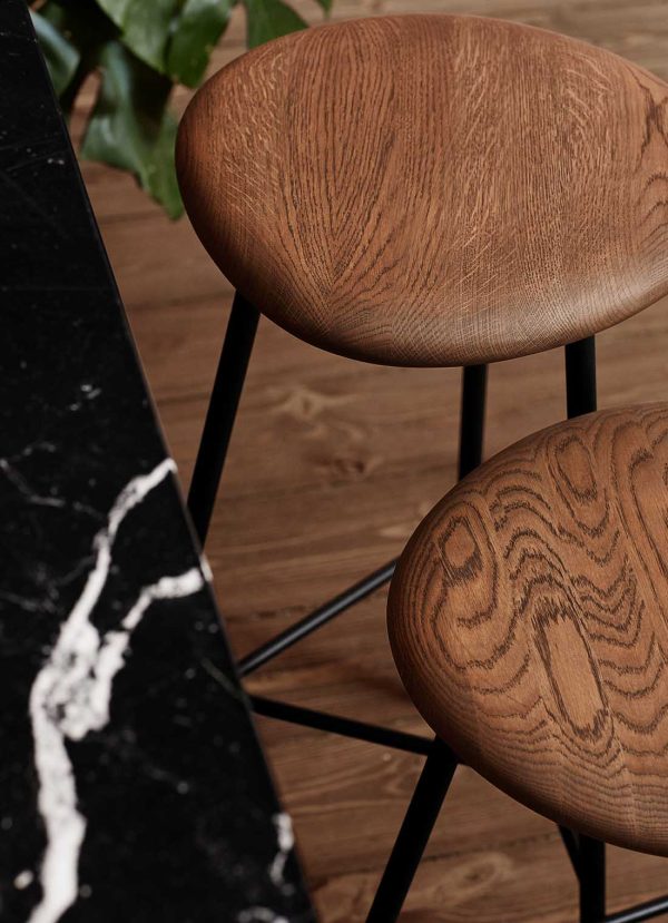 To moderne taburetter med afrundede træsæder og slanke sorte metalben, placeret ved siden af et elegant sort marmorbord med hvide årer, eksemplificerer banebrydende design. Baggrunden indeholder et strejf af grønt fra en plante.