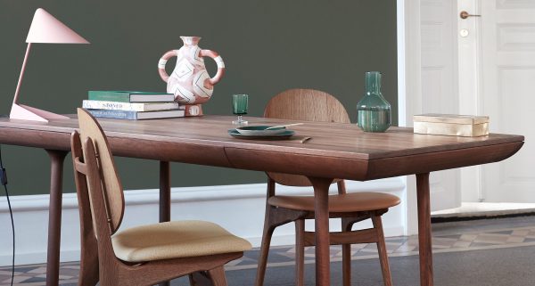 EN VARM NORDISK - Rúna Bord med stole fra midten af århundredet i et rum med olivengrønne vægge. på bordet står en lyserød lampe, dekorativt keramik, bøger, glas og tallerkener. en hvid dør er synlig i baggrunden.