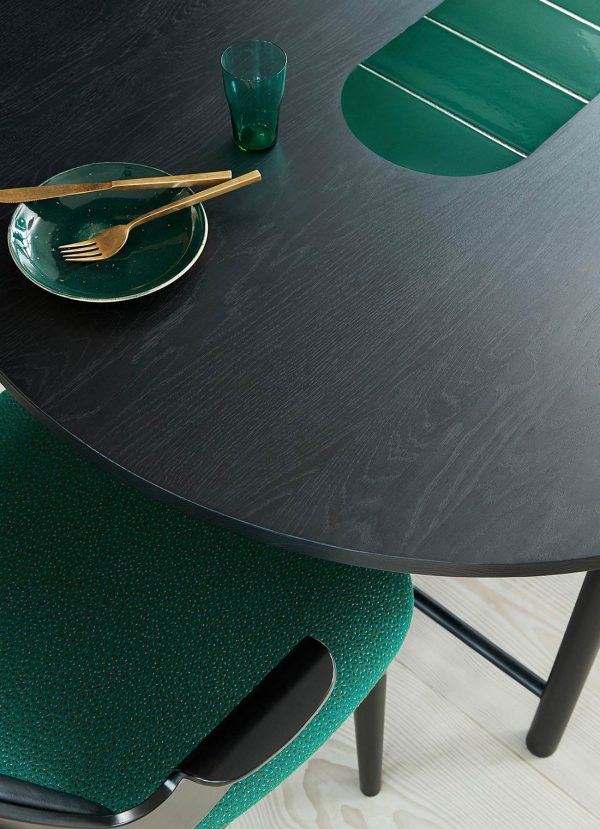 Et sort rundt bord med en grøn skål og glas, og gyldne redskaber på. Scenen inkluderer en grøn polstret designstol delvist skubbet under bordet.