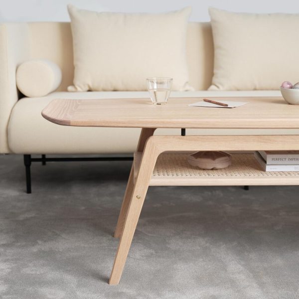 En moderne stue med et sofabord i træ med et glas på toppen og en notesblok med en blyant. En beige sofa med hynder og en vævet hylde under bordet er synlige. Blødt gråt tæppe designet af TomorrowsDesign dækker gulvet.
