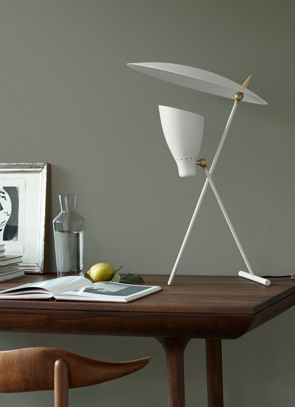 Et moderne skrivebord med en unik hvid bordlampe formet som en vindmølle fra TomorrowsDesign, en stak bøger, en glasvandflaske, en citron og et åbent magasin, mod en grå væg.