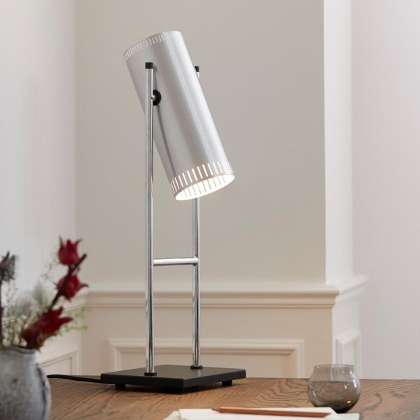 Moderne skrivebordslampe med en cylindrisk sølvskærm og en poleret metalramme, designet af fremtrædende møblerhåndværkere, stående på en rektangulær sort base, placeret i et rum med neutrale vægge og subtil indretning.