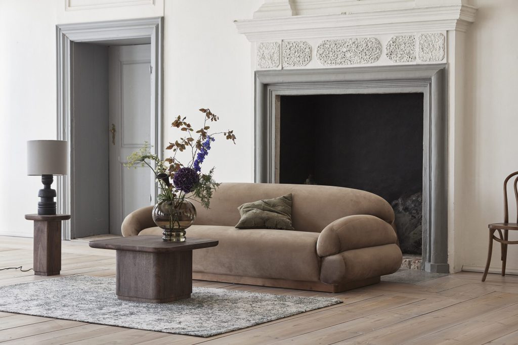 En stilfuld stue med en stor, udsmykket pejs, en plys beige sofa med hynder, designmøbler sofabord i træ og livlige blomster i vaser. Der er en lampe til venstre og en træstol til højre.