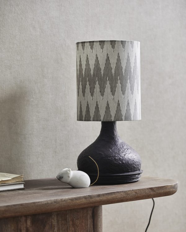 En tekstureret sort bordlampe med en ikat-mønstret skærm på et konsolbord af træ ved siden af en lille hvid figur og delvist synlige bøger, der inkarnerer essensen af TomorrowsDesign.