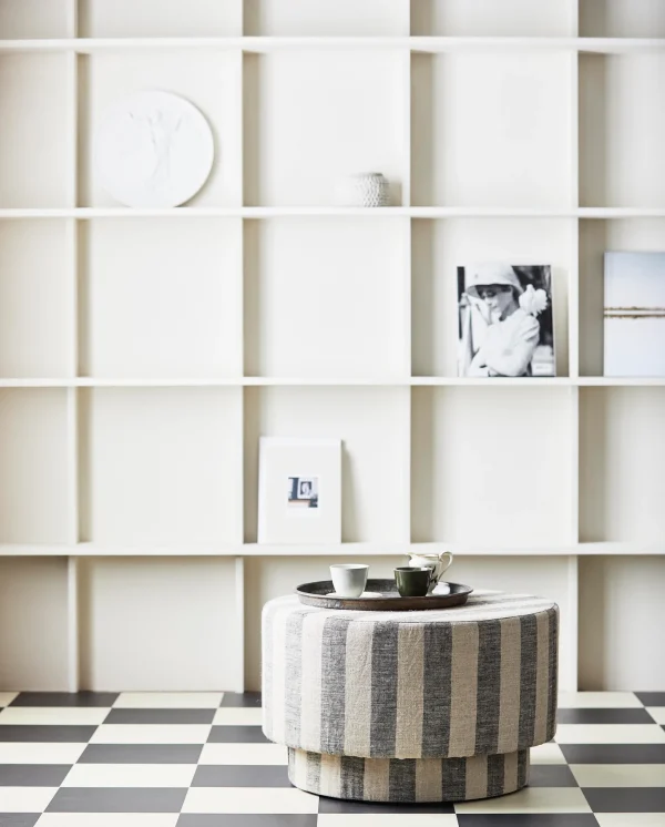 En moderne, minimalistisk stue med en hvid firkantet reol med et par dekorative genstande, herunder indrammede fotografier og en lille skulptur. En rund, stribet skammel med to kaffekopper sidder i forgrunden på et skakternet gulv, stylet af TomorrowsDesign.