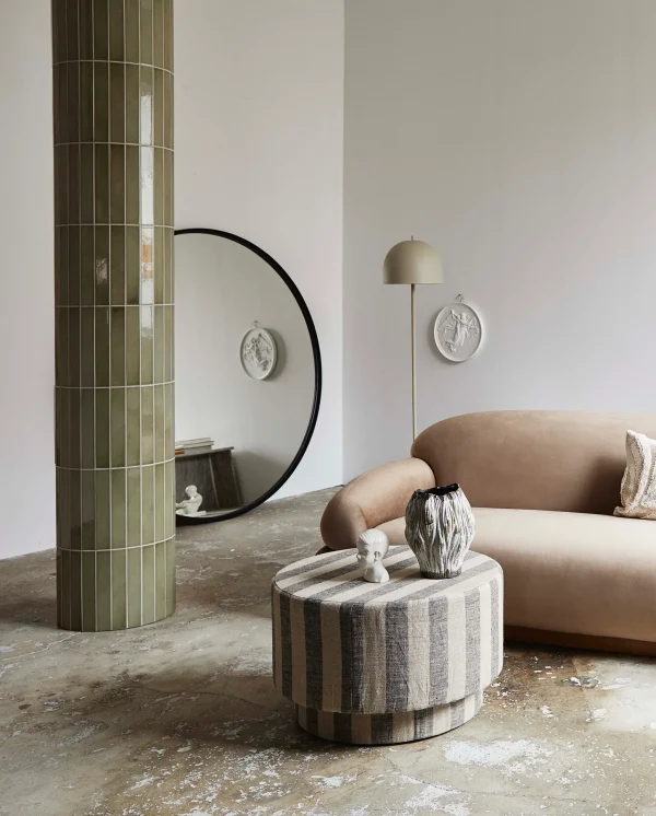 En minimalistisk stue med en plys beige sofa, en stribet rund skammel og en høj grøn flisebelagt søjle. Et stort rundt spejl afspejler en lille skulptur på skammel og dekorative plader på væggen. Rummet er et eksempel på elegant interiørdesign.