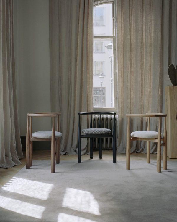 Et fredfyldt værelse med tre minimalistiske designstole, to af træ og en sort, placeret nær et draperet vindue, der kaster blødt lys og skygger på et plys tæppe.