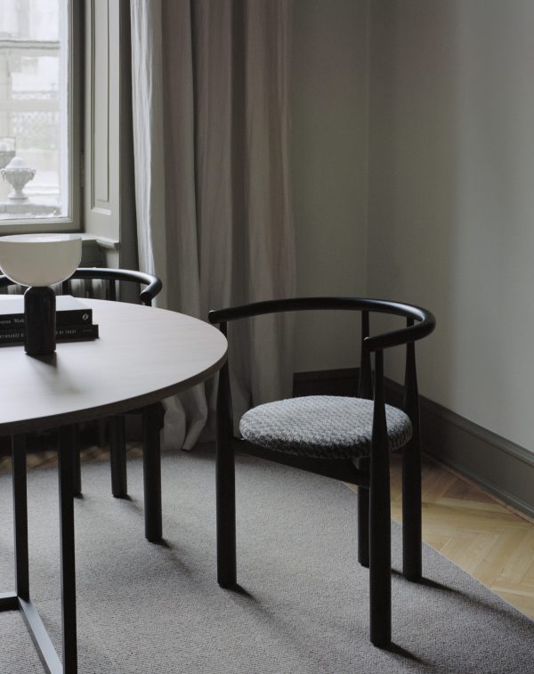 Et minimalistisk værelse med et rundt, hvidt bord og en sort stol med et vævet sæde. Et stort vindue med gennemsigtige gardiner giver naturligt lys, der komplementerer rummets neutrale farvepalet og interiørdesign.