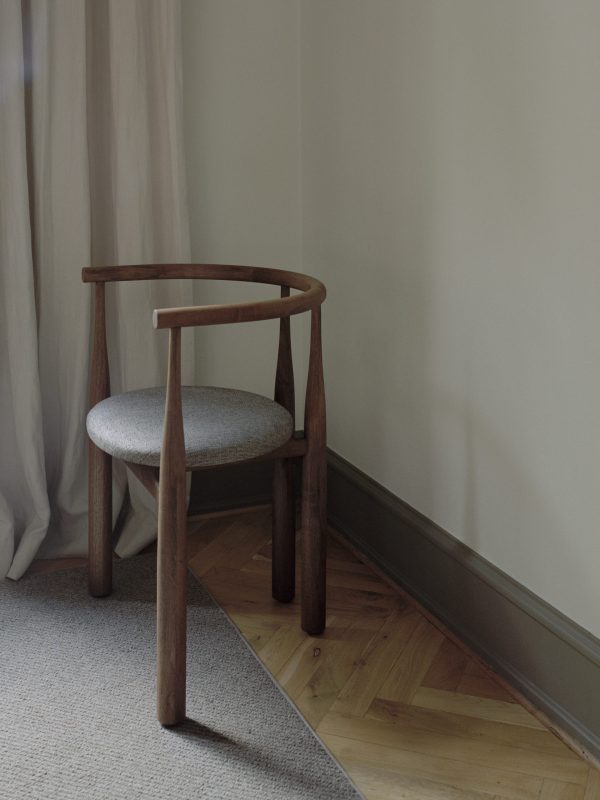 En træstol med buet ryglæn og gråt polstret sæde, placeret i et hjørne nær et vindue med gennemsigtige gardiner. Rummet har beige vægge og et sildebenstrægulv delvist dækket af et gråt tæppe, hvilket afspejler en elegant TomorrowsDesign-æstetik.